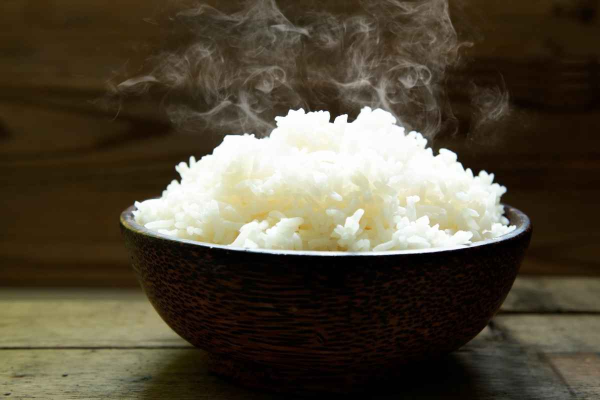 A dark wooden bowl filled with steaming white rice sits on a rustic wooden surface, with steam rising visibly from the rice.