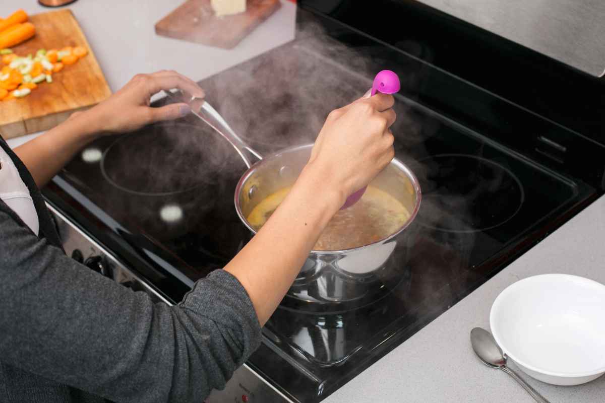 Person stirring soup in a pot on a stovetop with visible steam, a cutting board with chopped vegetables nearby, and an empty bowl on the counter.