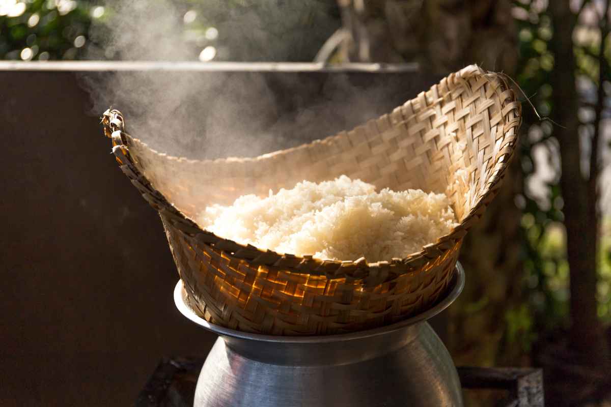A bamboo basket filled with steaming cooked rice sits on top of a metal pot, with steam rising in an outdoor setting.