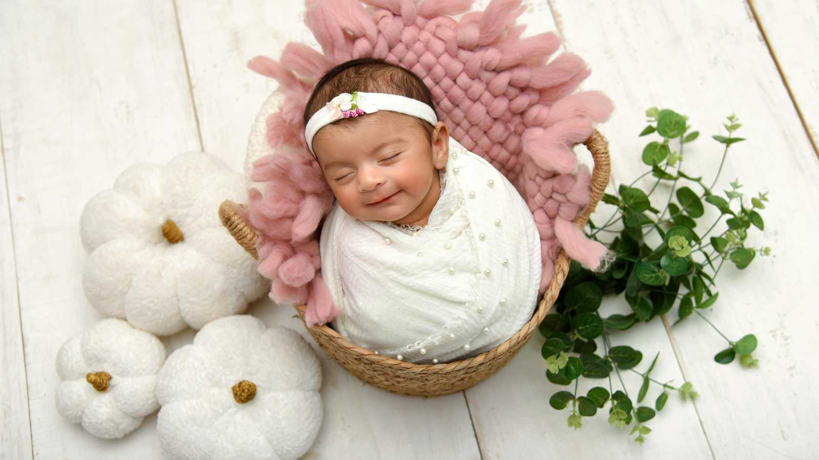 A newborn baby wrapped in a white blanket sleeps in a wicker basket decorated with pink fabric, next to white pumpkins and green foliage on a white wooden surface.