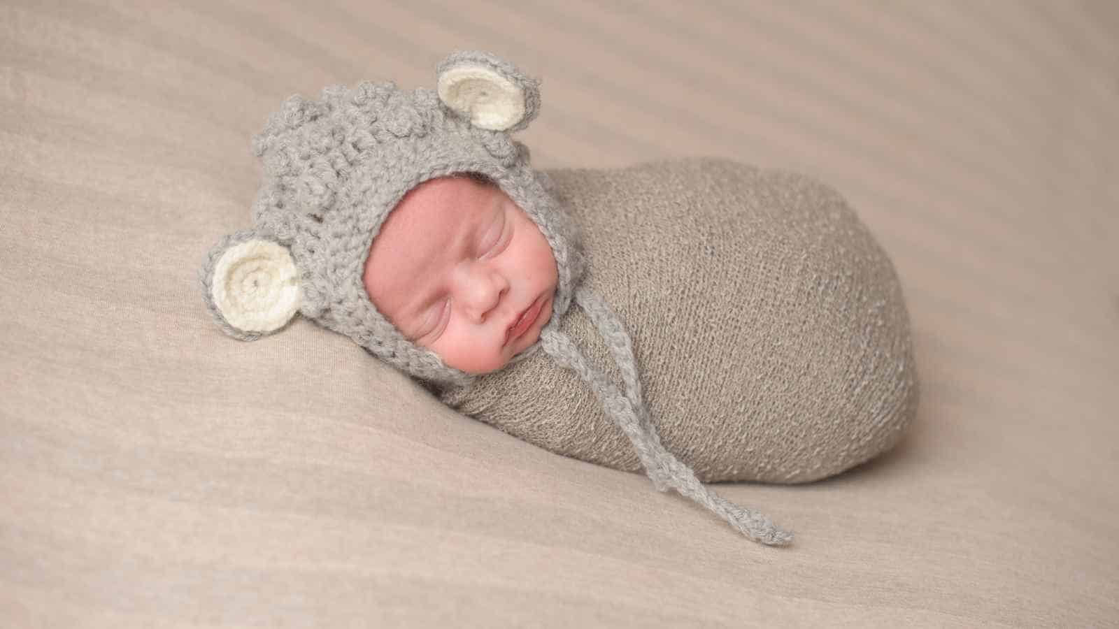 Newborn baby swaddled in a beige blanket, sleeping on a matching surface, wearing a knitted hat with bear ears.