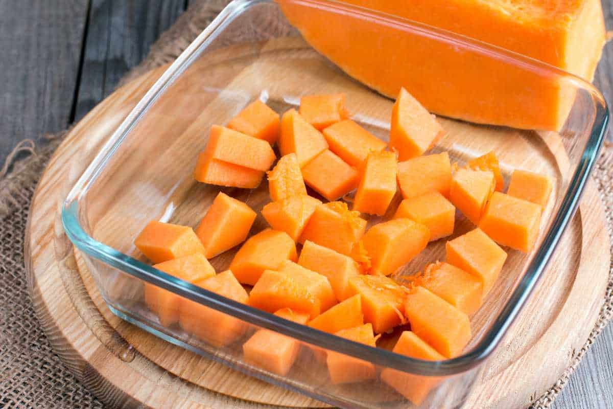 Glass dish with cubed raw pumpkin placed on a wooden board, with a larger piece of pumpkin in the background.