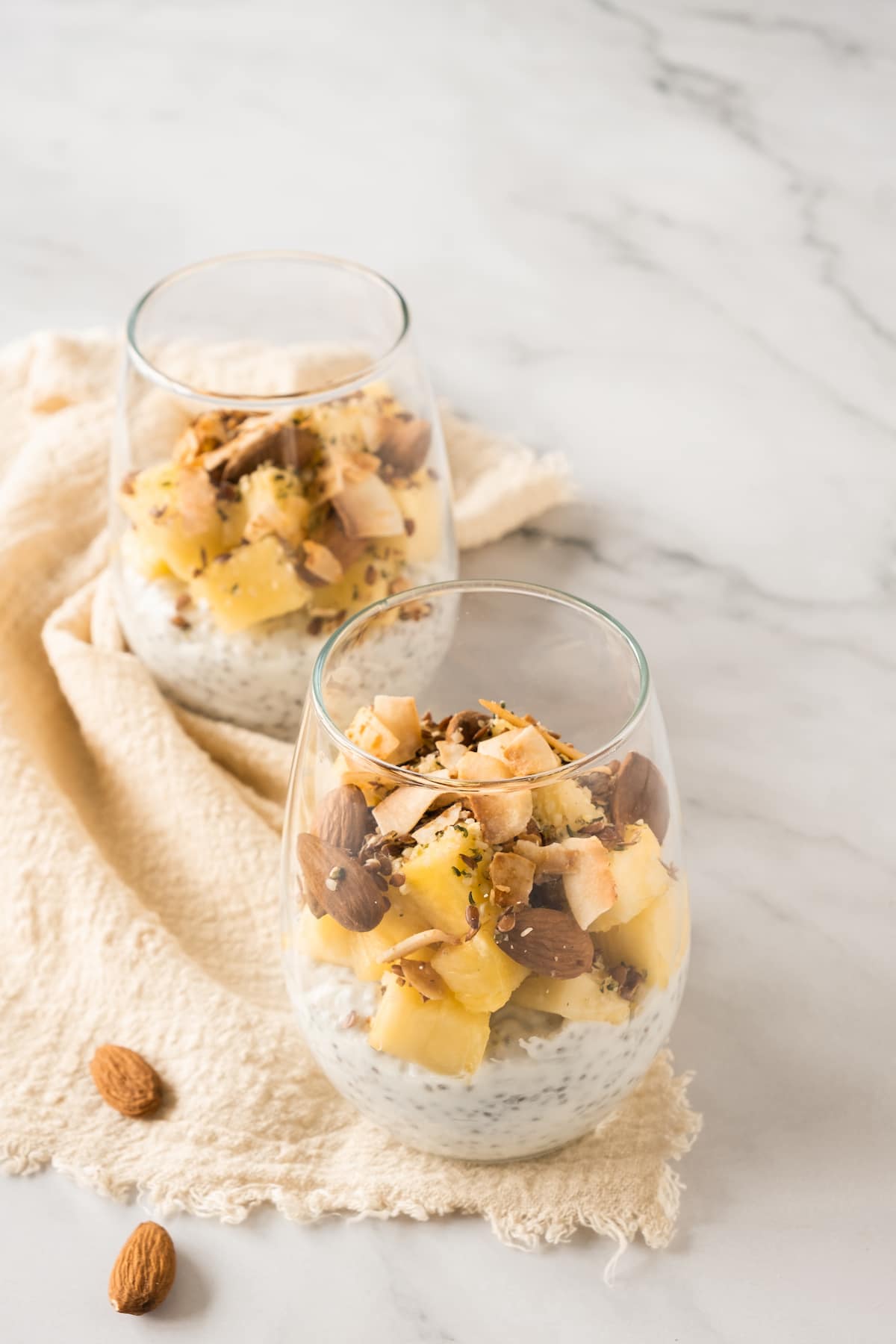 Two clear glasses filled with chia pudding, topped with pineapple chunks, almonds, coconut flakes, placed on a beige cloth on a marble surface.