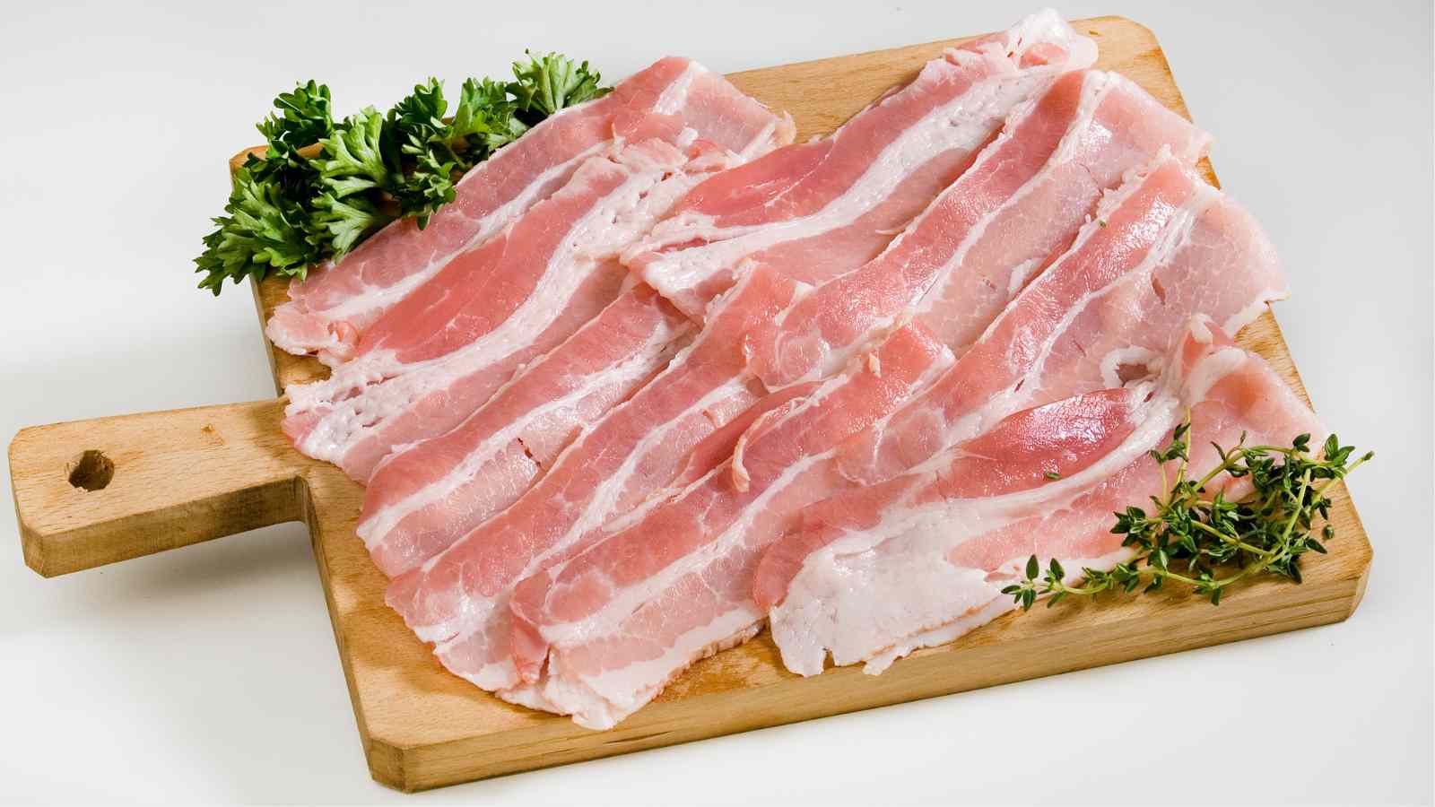 Several raw bacon strips are arranged on a wooden cutting board, garnished with sprigs of fresh parsley and thyme.