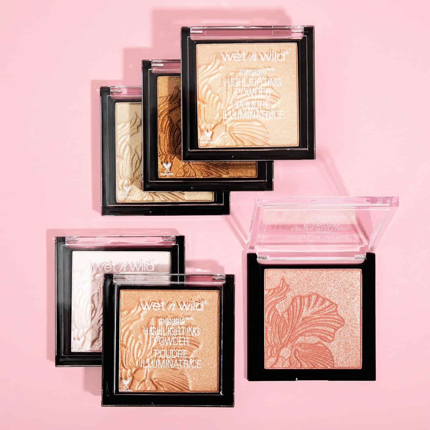 Six Wet n Wild highlighter compacts in various shades are arranged on a pink surface, some with lids open to show embossed powder designs.