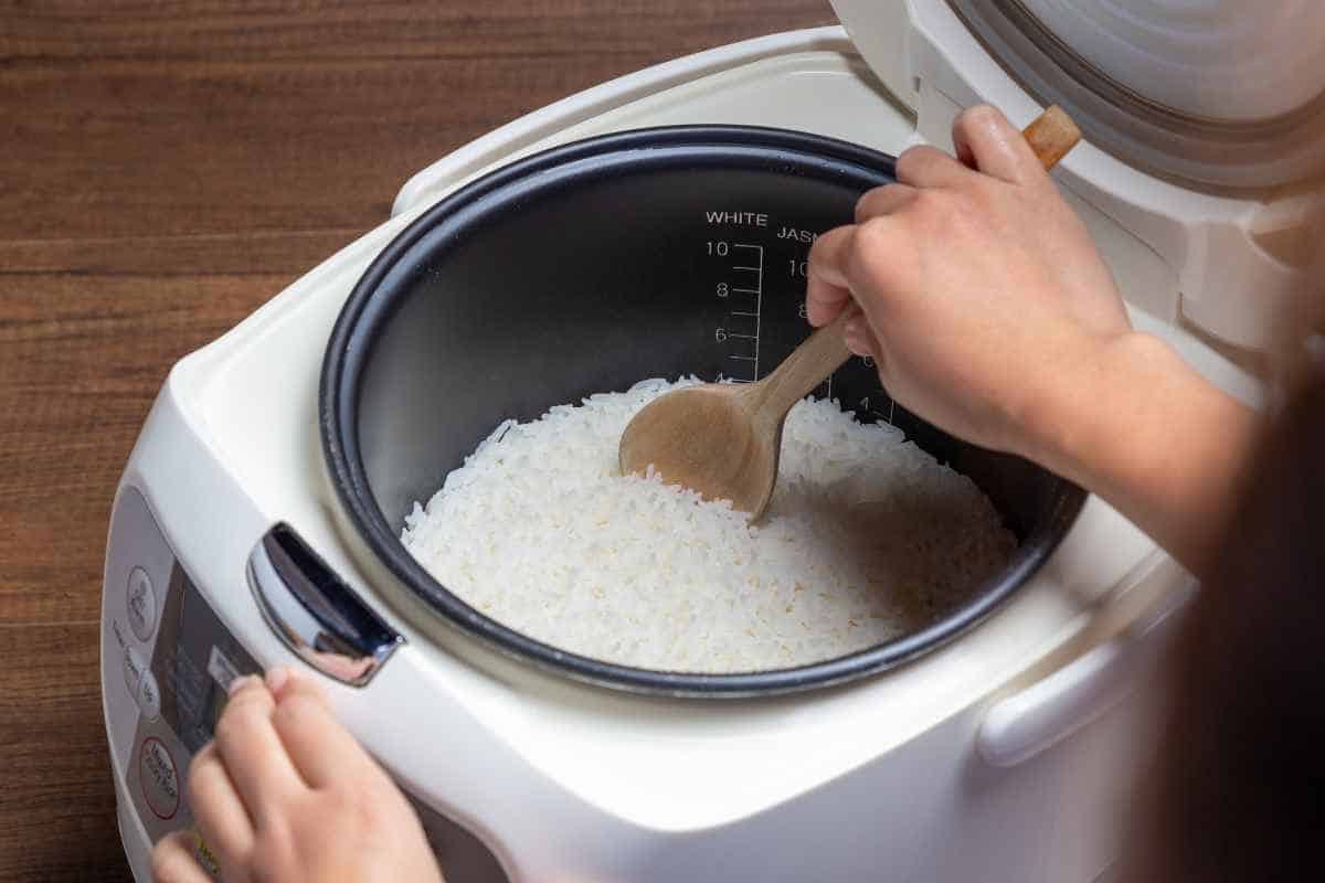 A person stirs cooked white rice in a rice cooker using a wooden spoon.