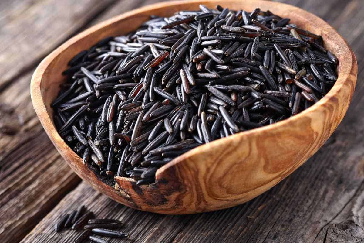 A wooden bowl filled with uncooked wild rice sits on a rustic wooden surface.