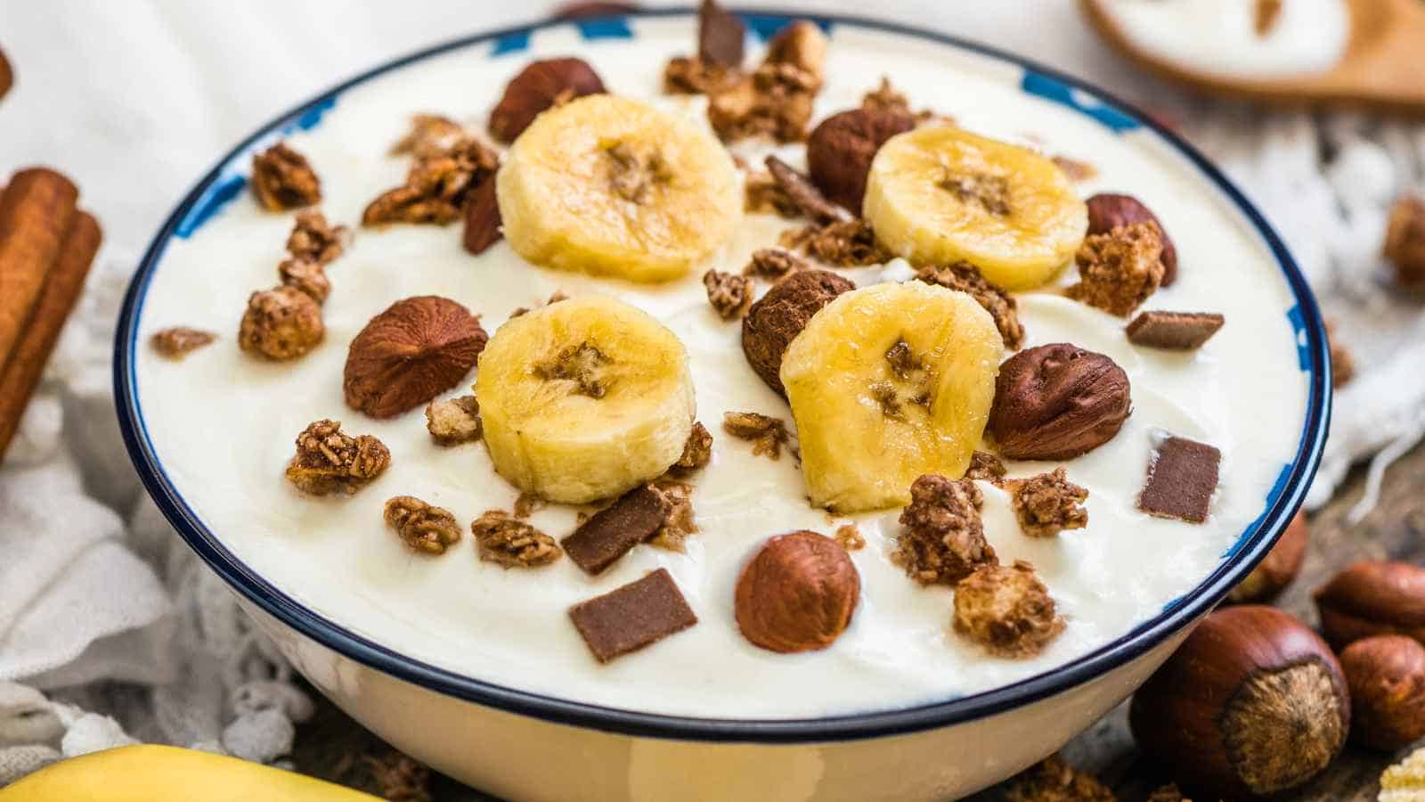 A bowl of yogurt topped with banana slices, granola, hazelnuts, and chocolate chunks.