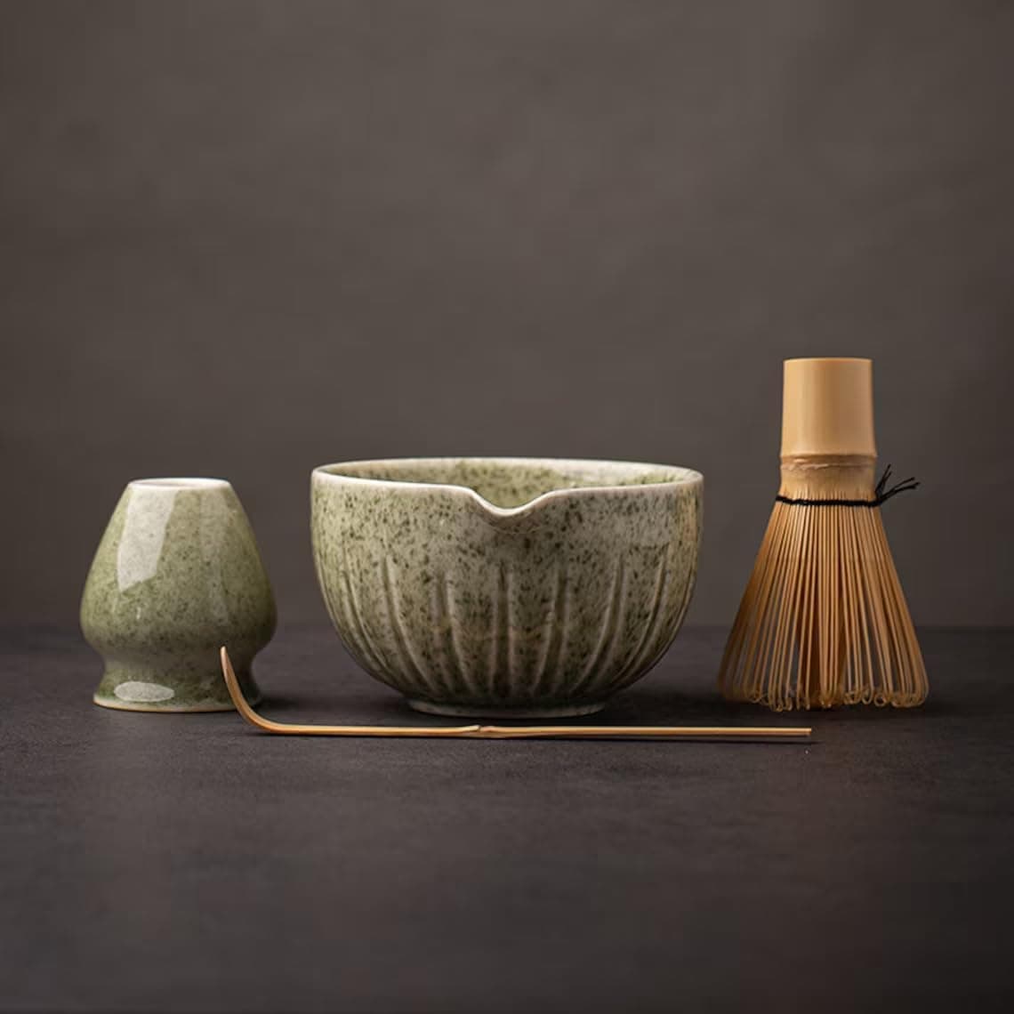 A matcha tea set with a whisk, a ceramic bowl, a small ceramic holder, and a bamboo scoop displayed on a dark surface.