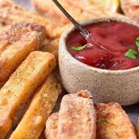 Thick-cut air fried halloumi fries arranged around a small bowl of ketchup with a spoon, garnished with parsley and lemon wedges, on a round plate.