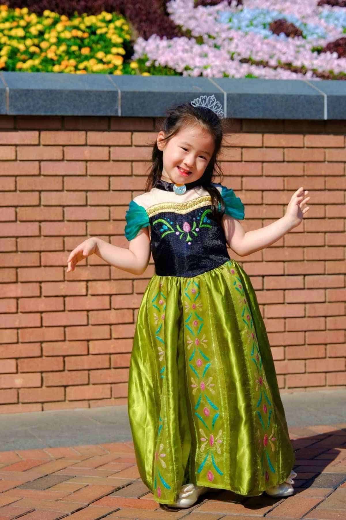 A young girl in a green and black princess dress with a tiara poses and smiles outdoors in front of a brick wall and flower bed.