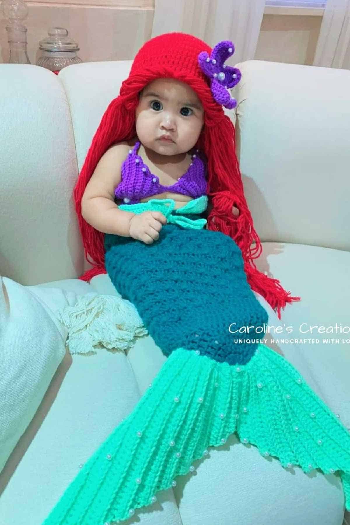 A baby dressed in a crocheted mermaid costume with a red wig, purple top, and teal tail sits on a white couch.