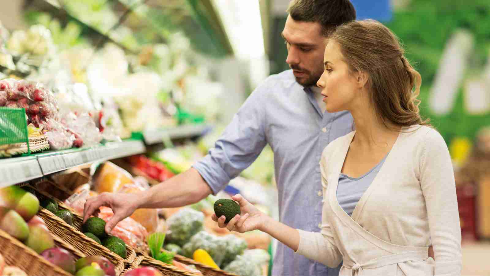 A couple shops for produce in a grocery store, examining avocados.