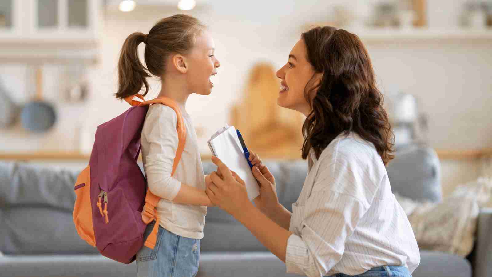 A mother helps her young daughter get ready for school by putting her books and a pen into her backpack.