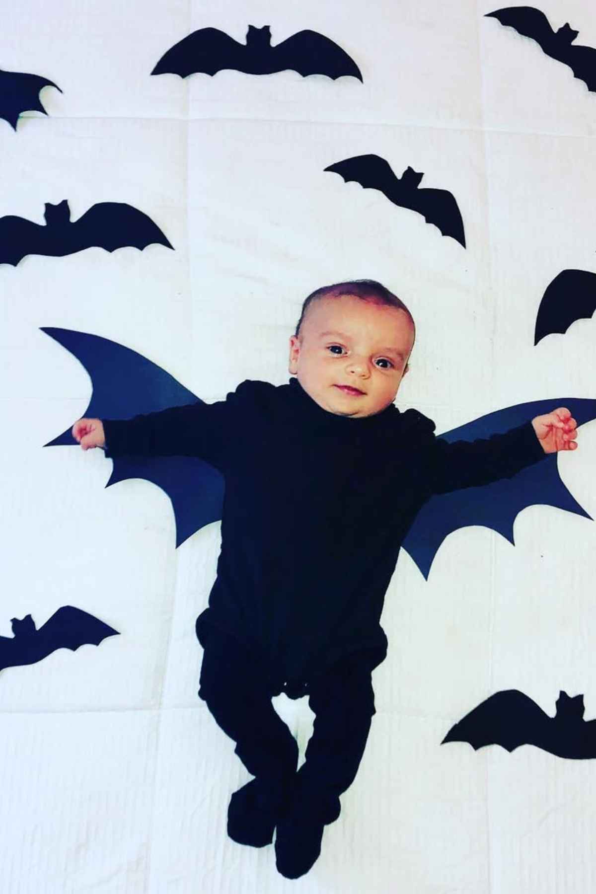 A baby wearing all black onesie and a bat wings while lying on bed. Bat papers scattered on his side.