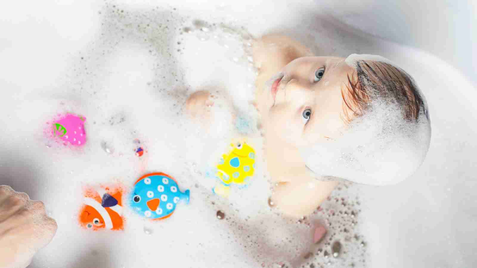 A baby with bubbles on their head sits in a bathtub surrounded by colorful toy fish, looking upwards.