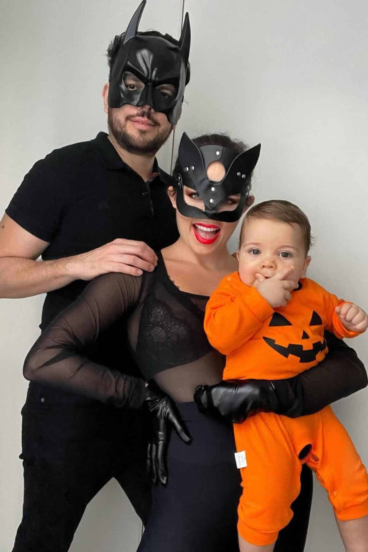 Three people pose in costumes: an adult in a Batman mask, another adult in a Catwoman mask, and a baby in an orange pumpkin outfit.