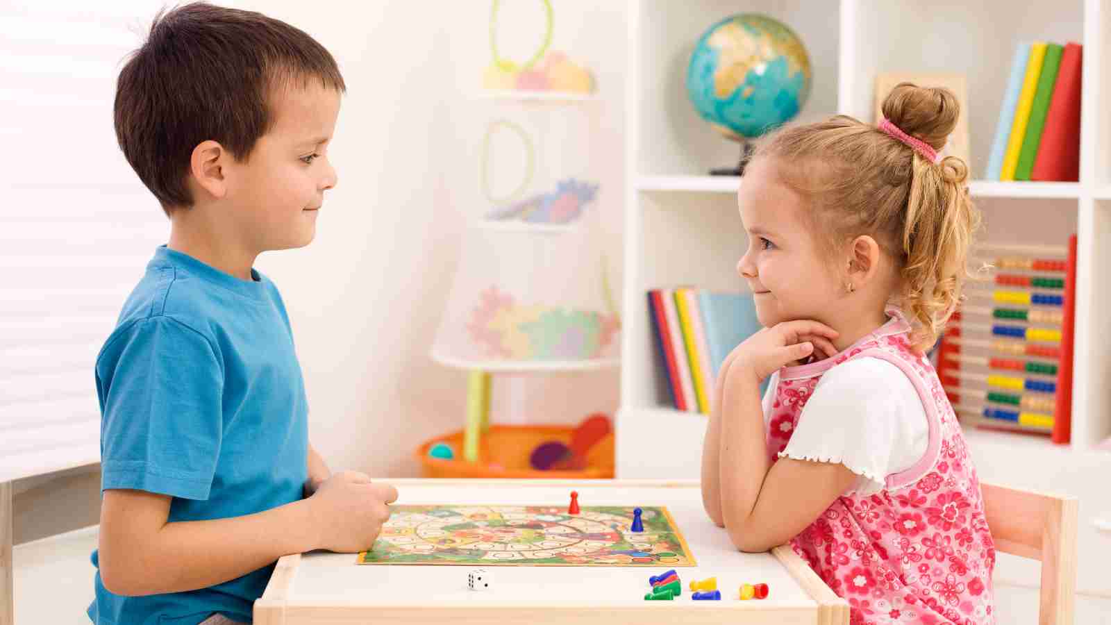 Two children sit at a small table playing a board game, facing each other and smiling, with shelves of books and toys in the background.