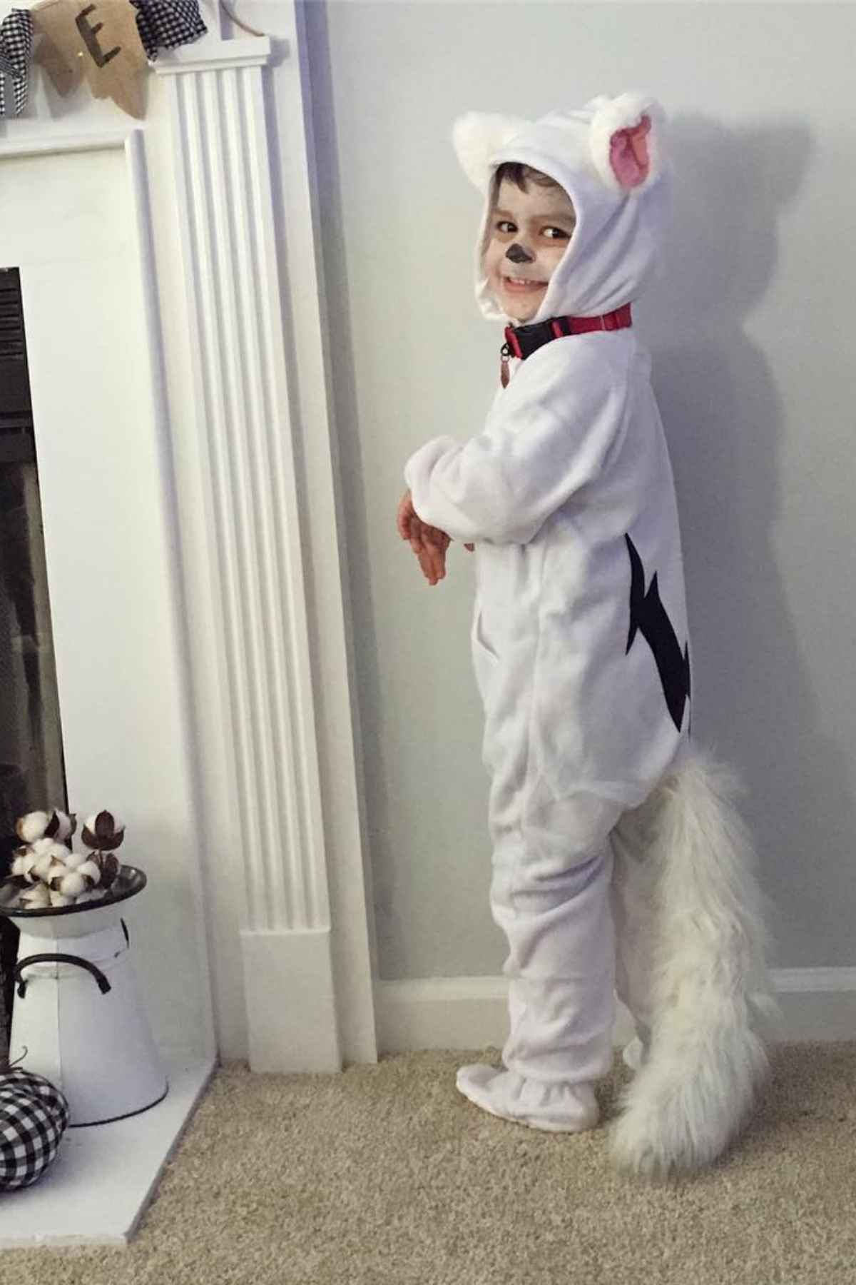 A child wearing a white dog costume with a tail, ears, and face paint stands indoors next to a fireplace.