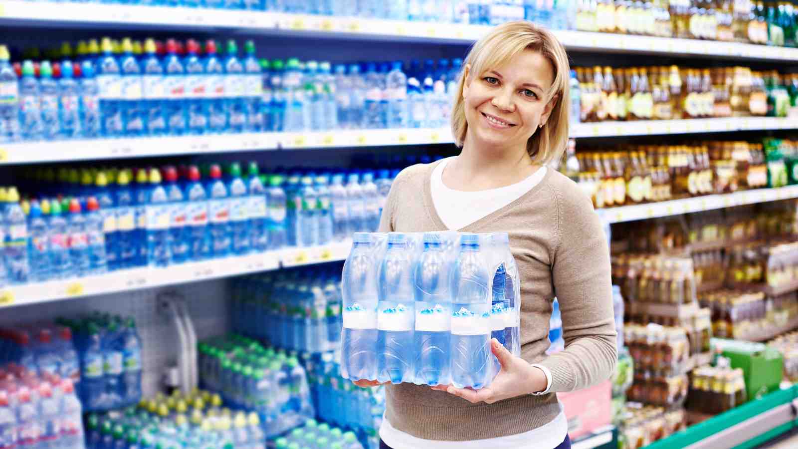A woman smiles at the camera while holding a multi-pack of bottled water in a supermarket aisle.