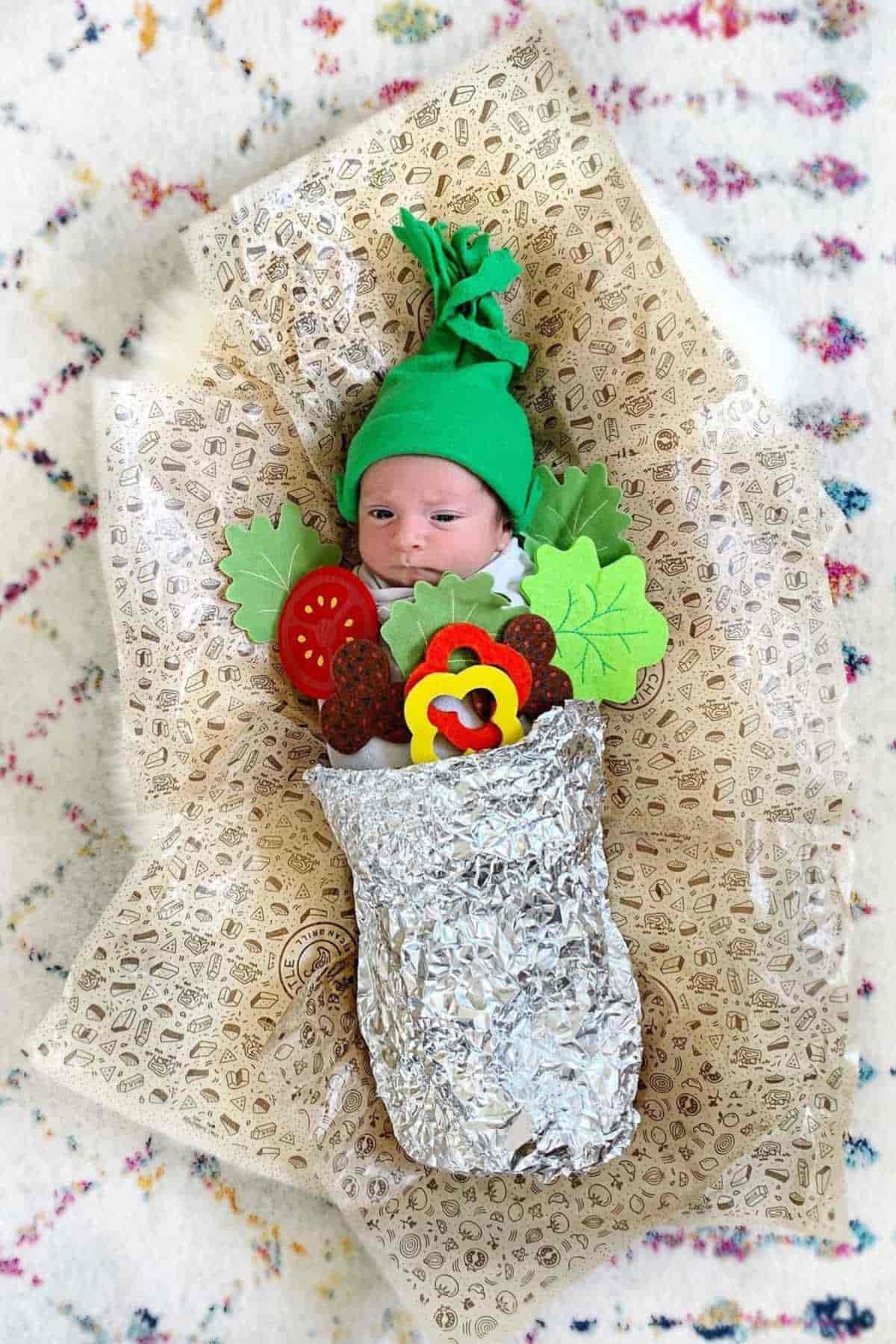 A baby wearing a green hat dressed as a burrito, lying on a foil and a wrapper.