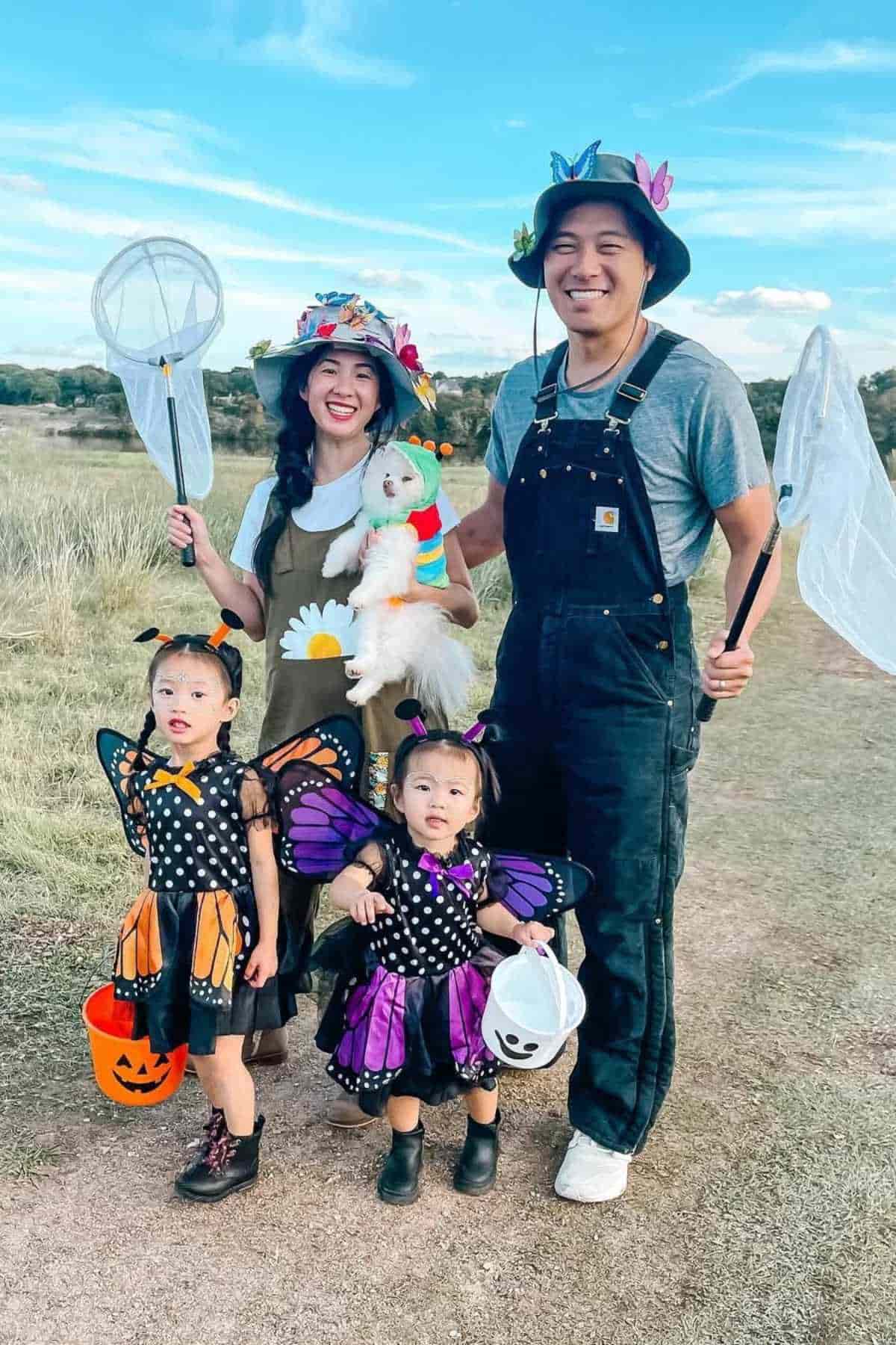 A family of four in a field wearing butterfly and flower costumes; two adults hold bug nets and two children are dressed as butterflies with Halloween buckets.