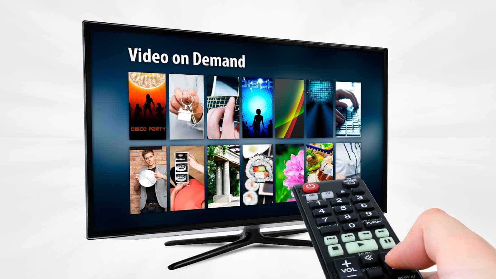 A hand holds a TV remote pointed at a flat-screen television displaying a video-on-demand menu.