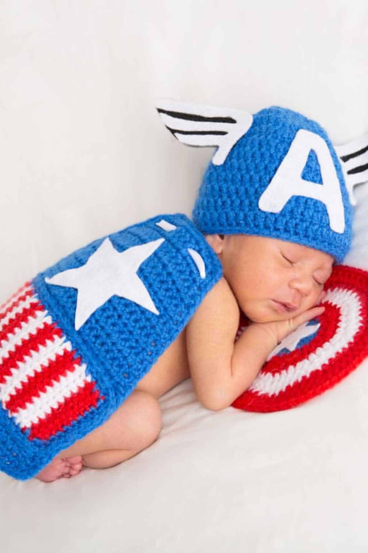 Baby dressed in a crochet captain America costume while lying on his back in bed.