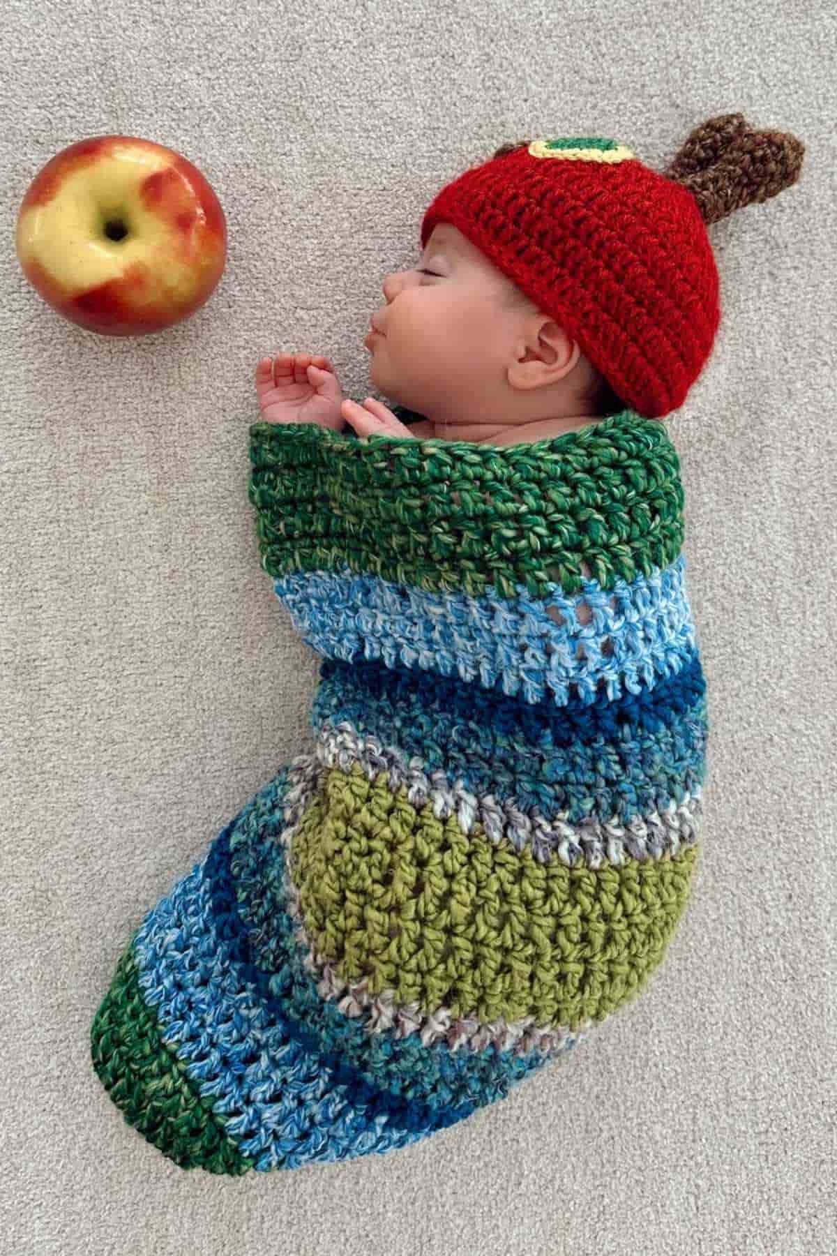 A baby is wearing a crochet caterpillar costume with a red hat, lying beside an apple.