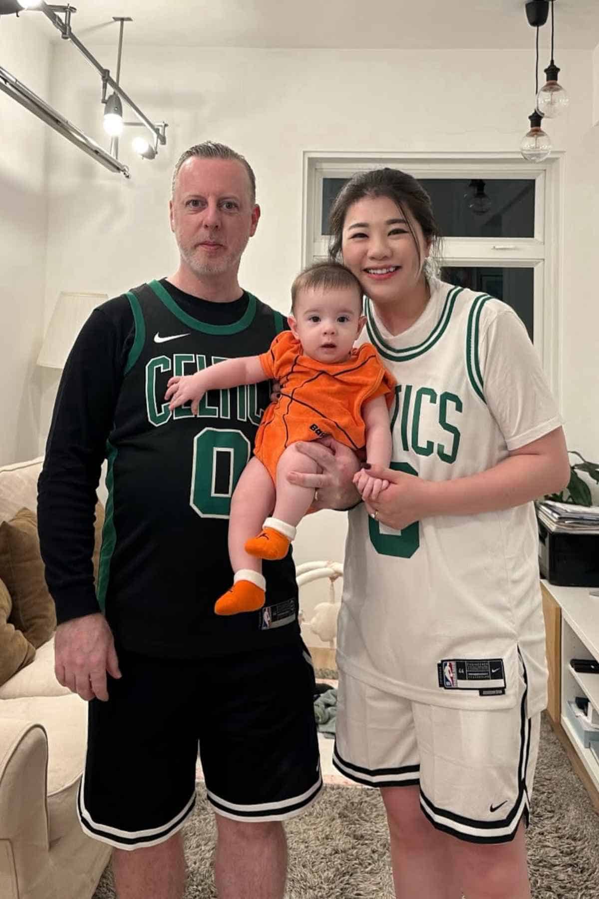 Two adults in Boston Celtics jerseys stand indoors with a baby dressed in an orange outfit; all are smiling and posing for the photo.