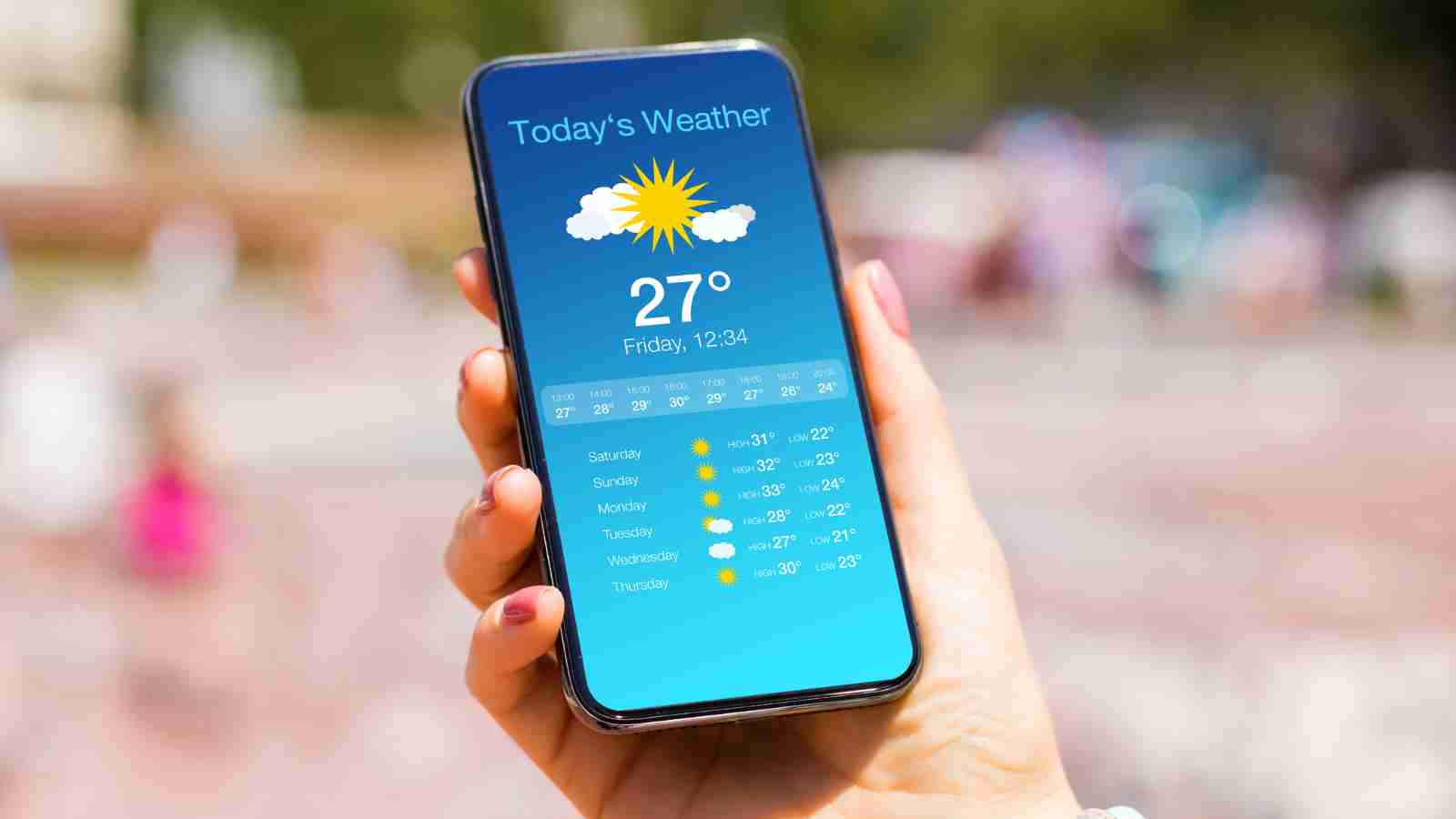 A person holds a smartphone displaying a weather app showing the current temperature and a five-day forecast.