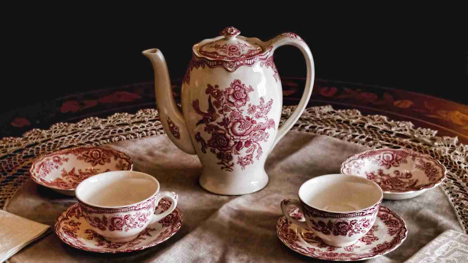 A teapot and two teacups and saucers with a red floral pattern sit on a lace tablecloth.