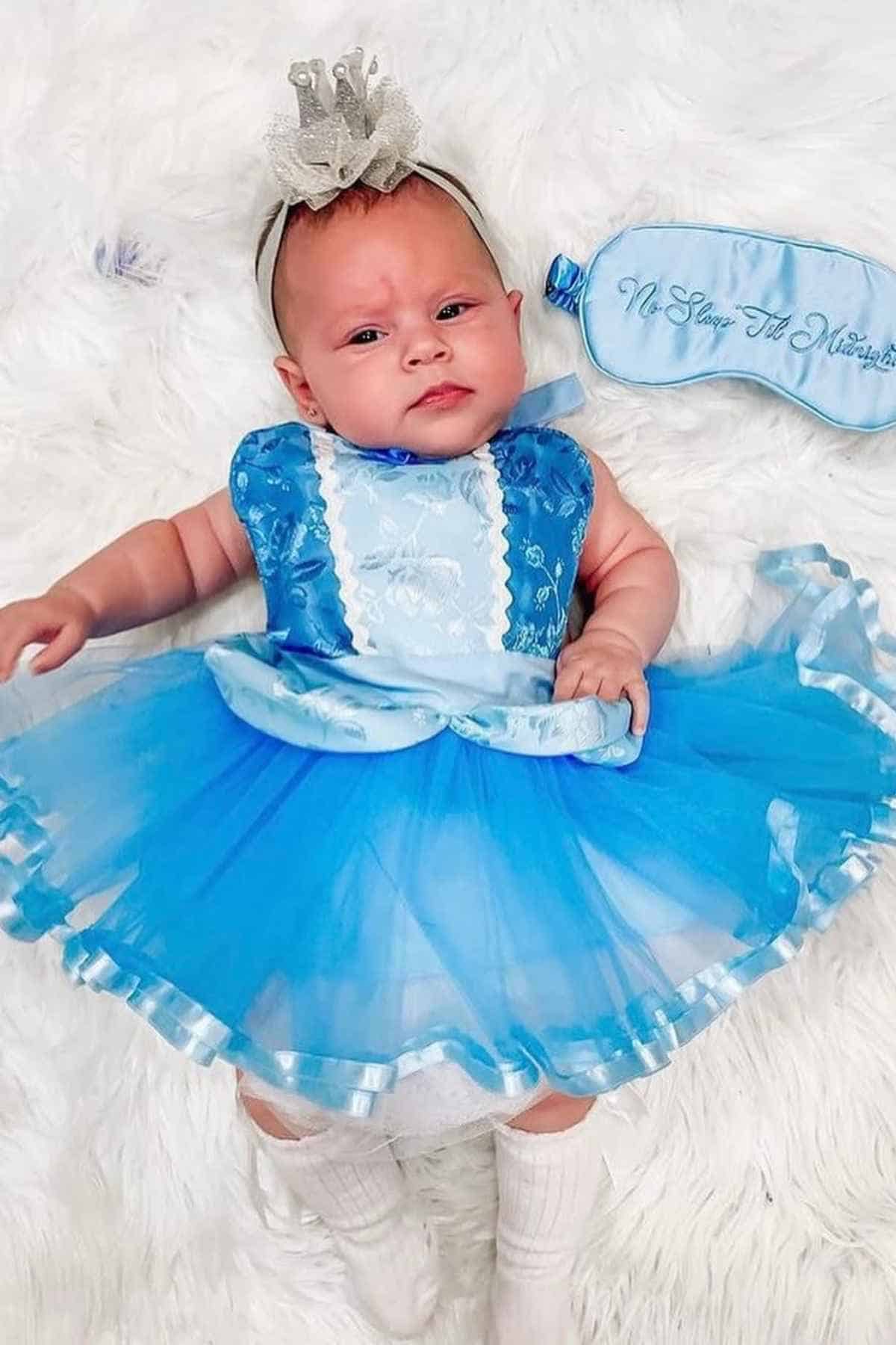 Baby wearing a blue dress, crown headband, and white socks lying on a white surface with a blue sleep mask beside her.