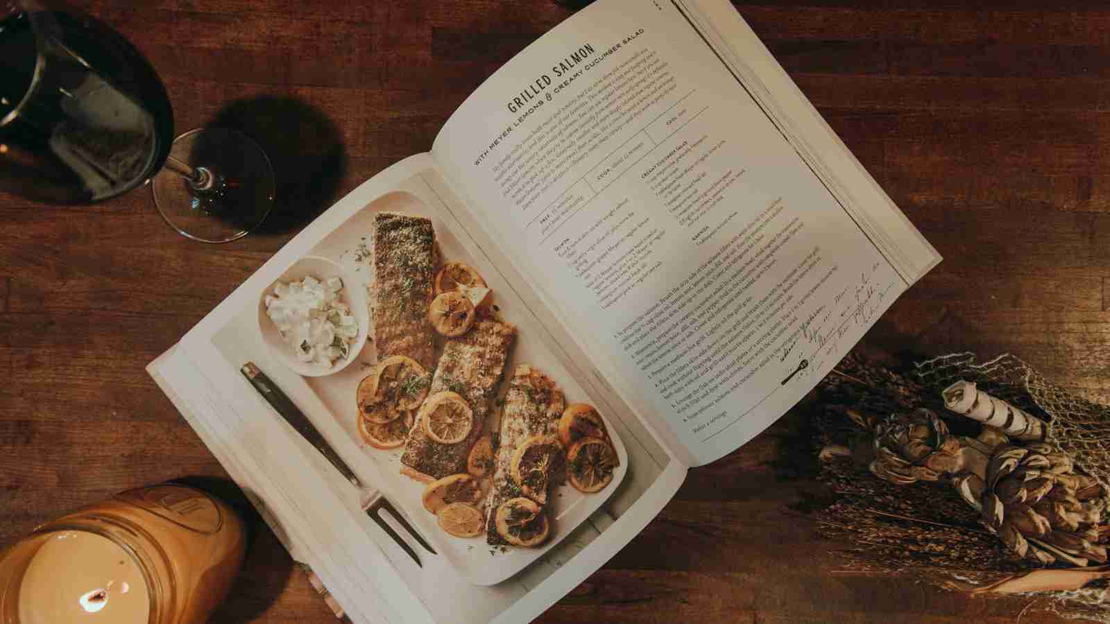 An open cookbook displaying a recipe for grilled salmon with lemons and a creamy cucumber salad is shown on a wooden table next to a glass of wine and a candle.