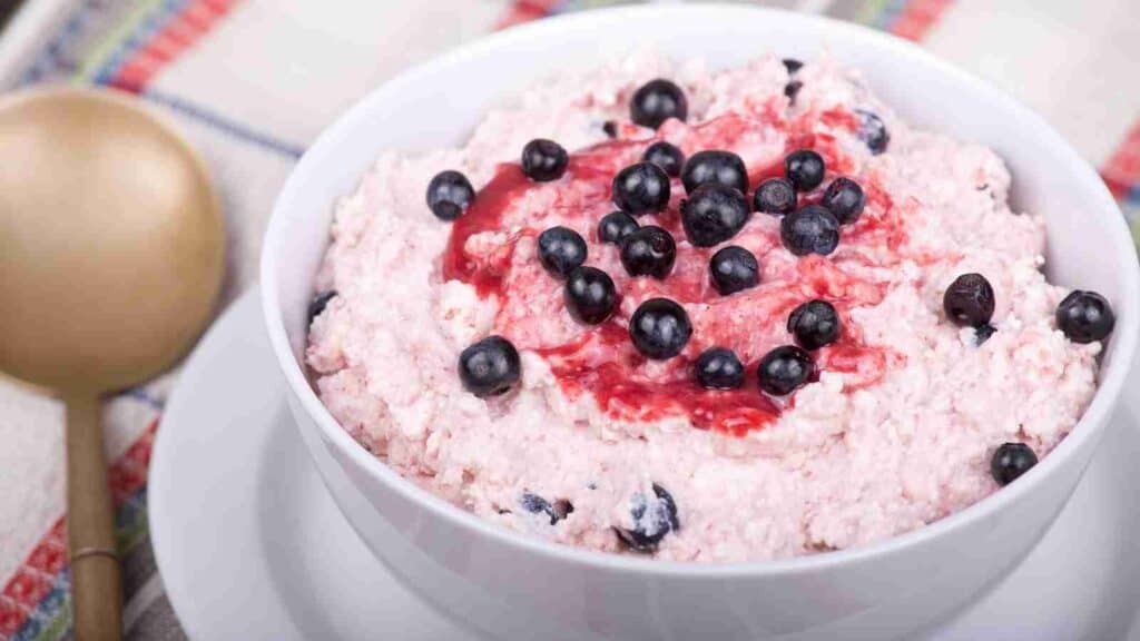A bowl of creamy pink dessert, topped with blueberries and a berry sauce, is presented on a white plate next to a wooden spoon.