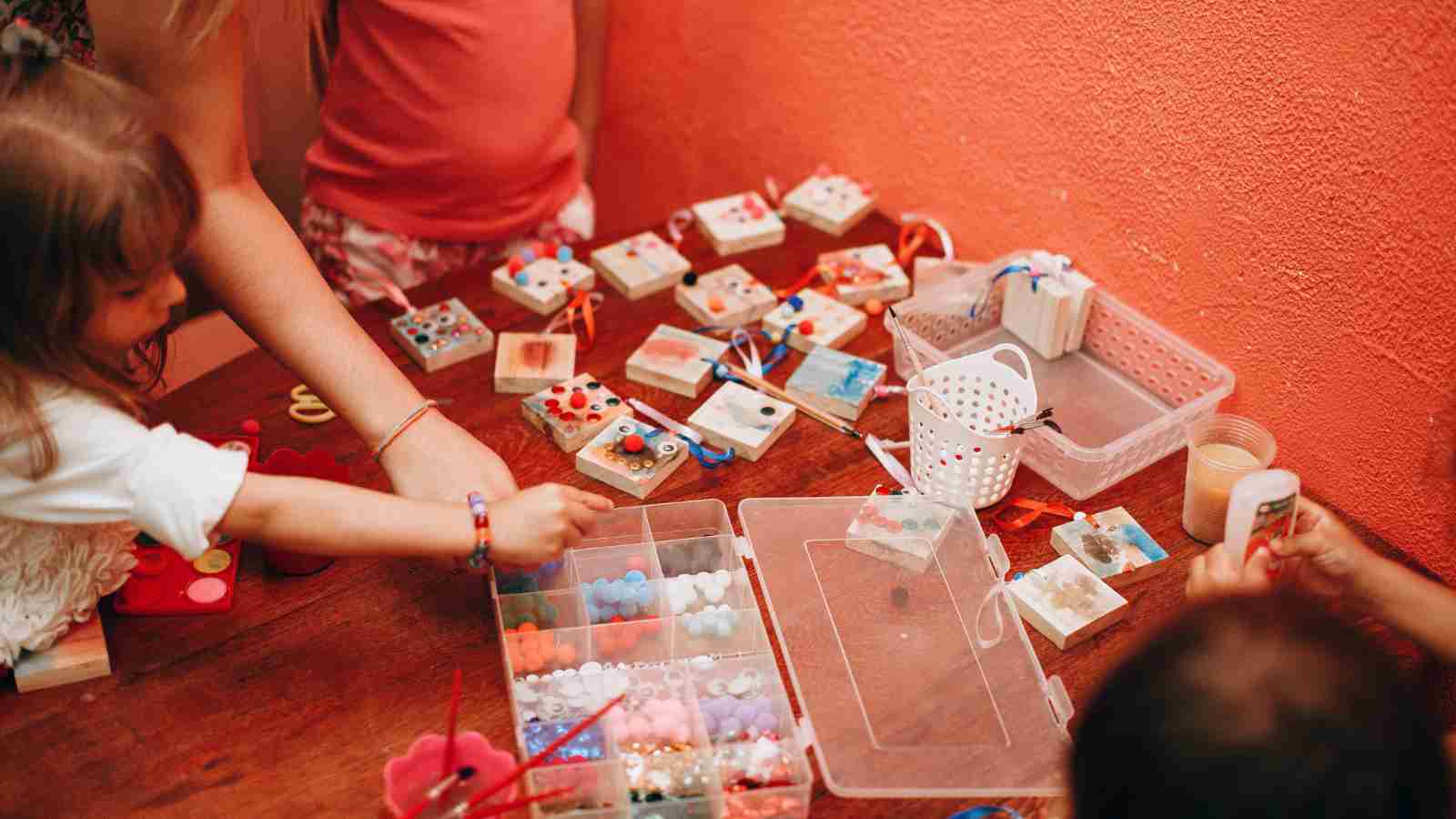 Children and an adult sit at a table, crafting with small colorful beads and decorated boxes, with various materials organized in plastic containers.