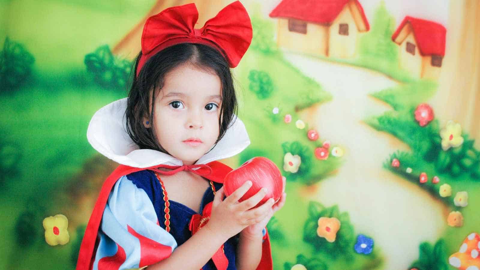 A young child in a Snow White costume holds a red apple, standing in front of a colorful, fairy-tale themed backdrop with houses and flowers.