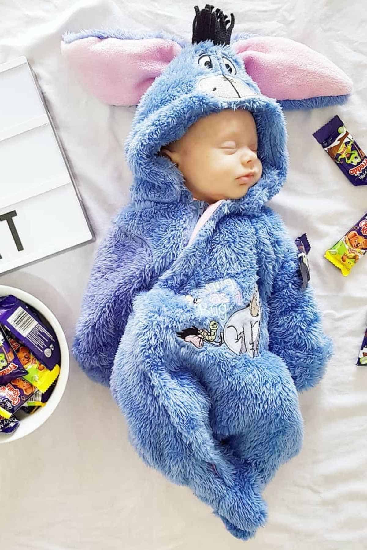 Baby dressed with a blue donkey costume while on bed, surrounded by some chocolates.