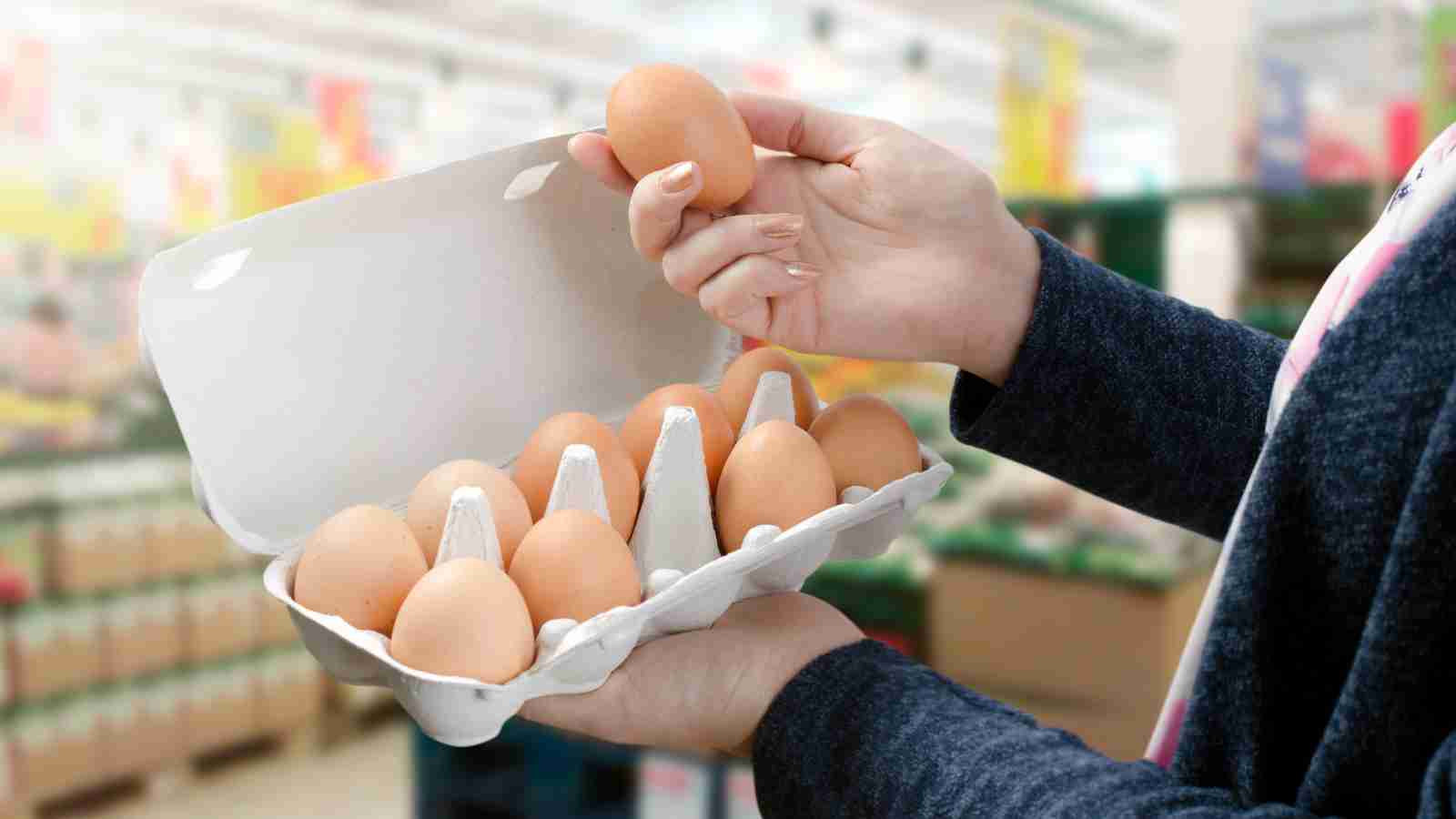A person's hands hold a carton of eggs in a grocery store, selecting one egg to examine.