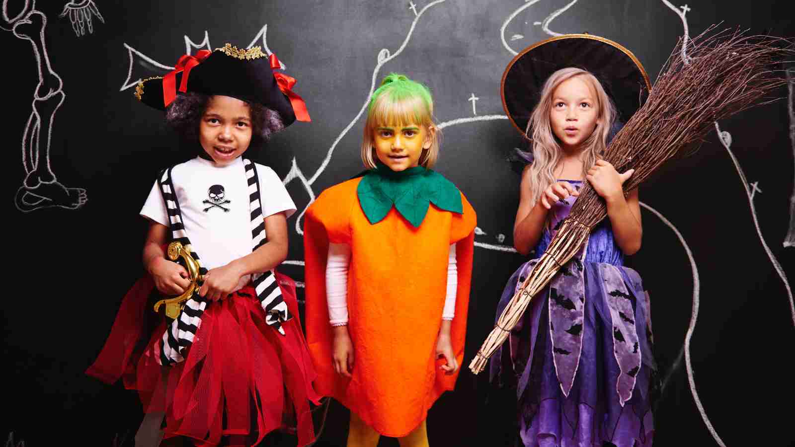 Three children in Halloween costumes stand in front of a chalkboard; one is a pirate, one a carrot, and one a witch holding a broom.