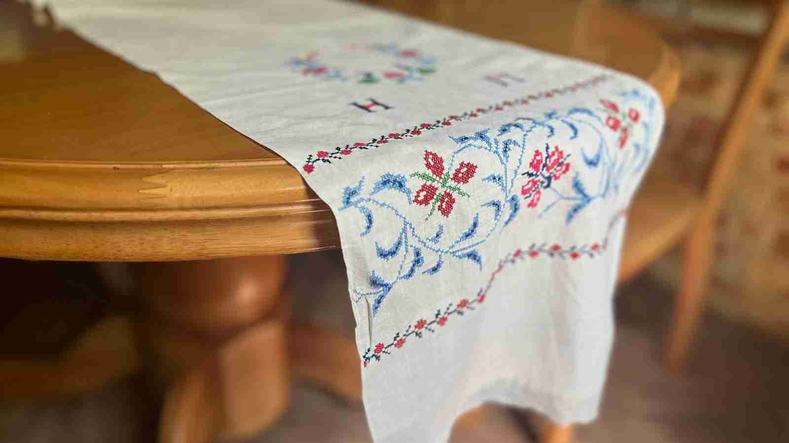 A white linen table runner with embroidered flowers and the letter "H" is draped over a wooden table.