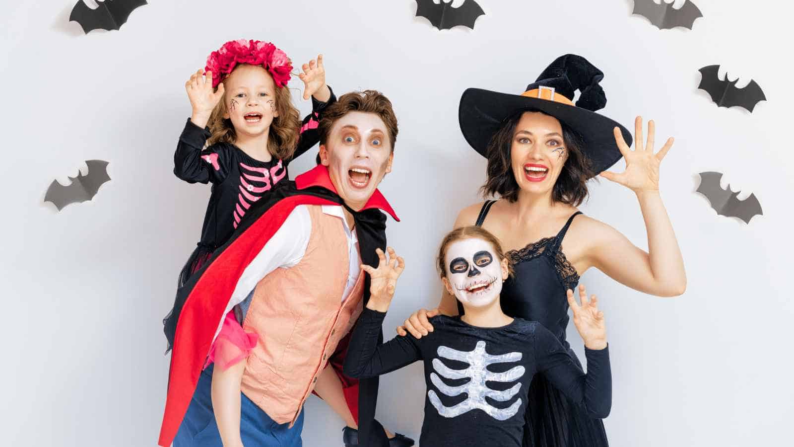 A family dressed in Halloween costumes poses in front of a wall decorated with paper bats; two children wear skeleton outfits, one adult is a witch, the other is a vampire.