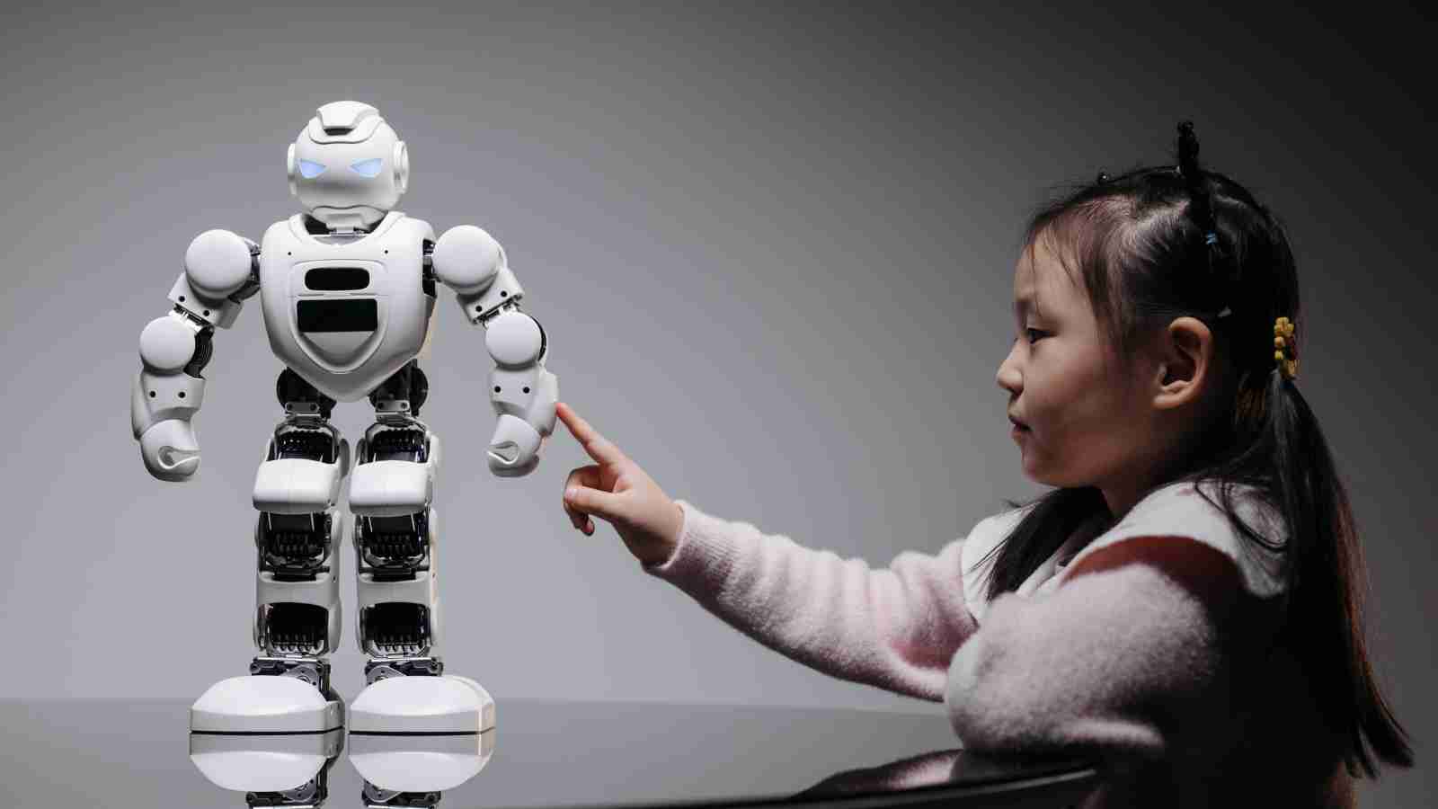 A young girl sits at a table and touches the arm of a humanoid robot standing beside her against a neutral background.
