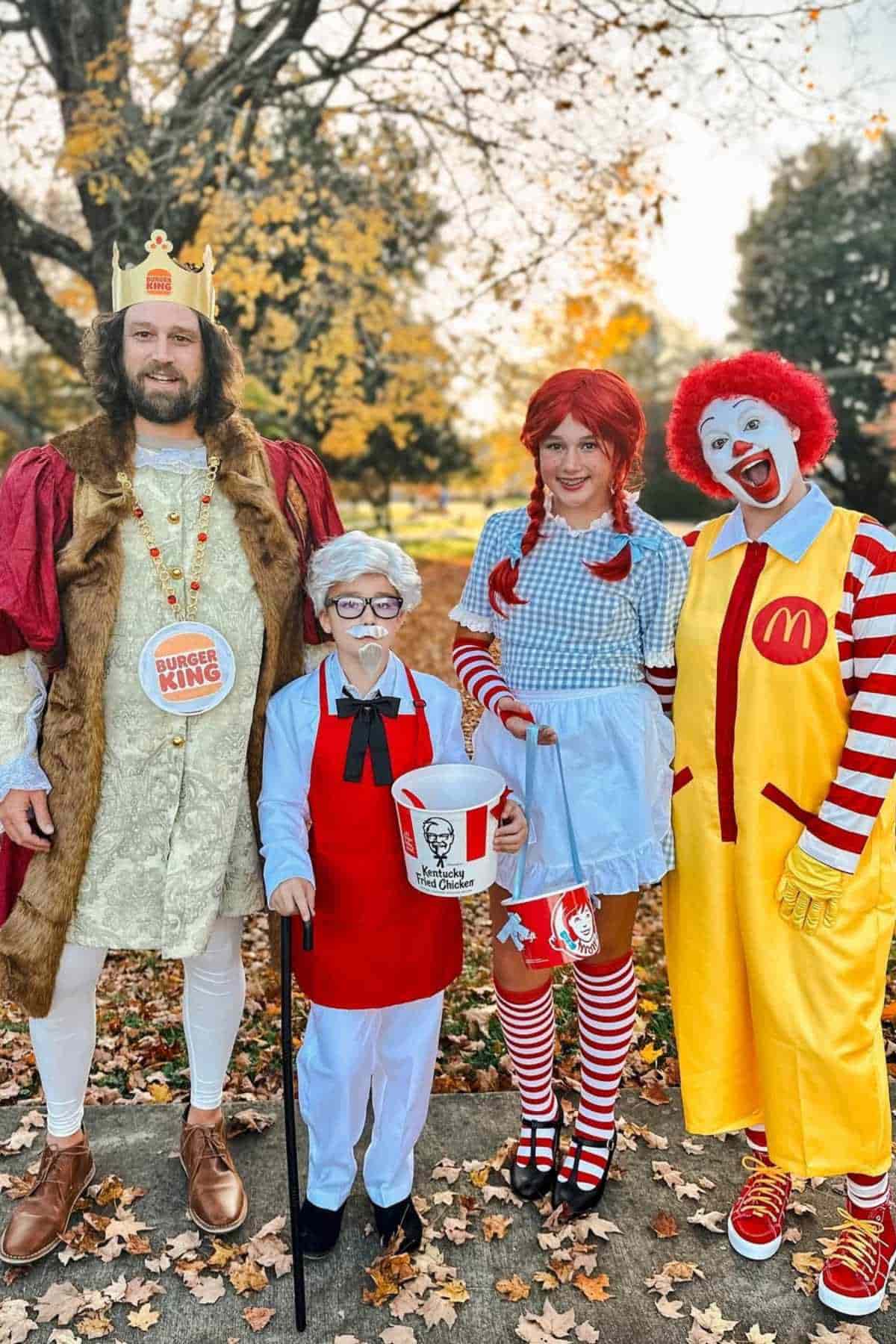 Four people dressed in fast food mascot costumes: Burger King, Colonel Sanders, Wendy, and McDonald, standing outdoors on a leaf-covered sidewalk.