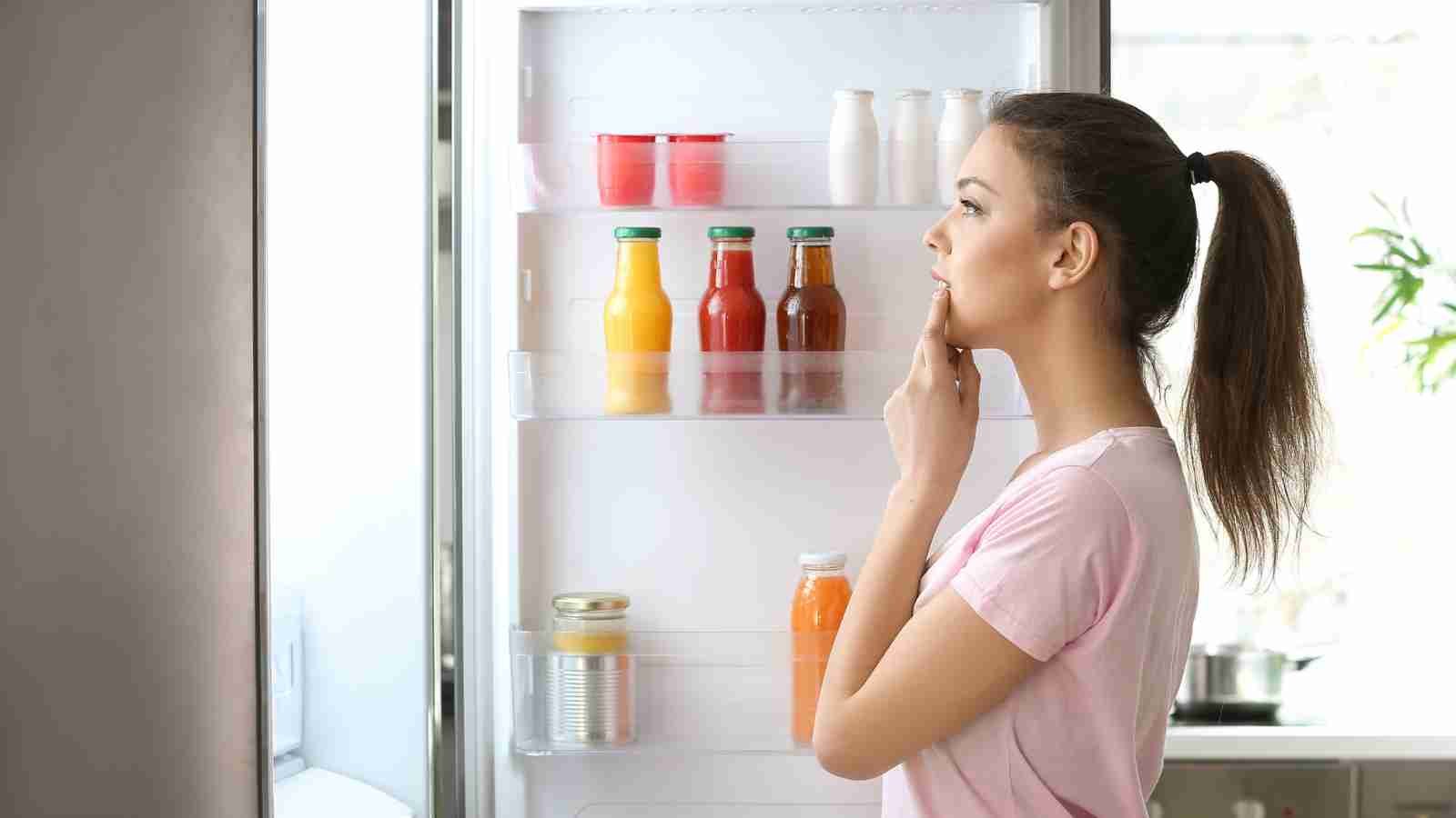 A woman stands thoughtfully in front of an open refrigerator, considering her food options.