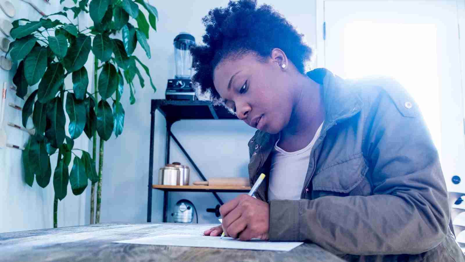 A young woman with dark curly hair sits at a table, concentrating on writing on a piece of paper with a pencil.