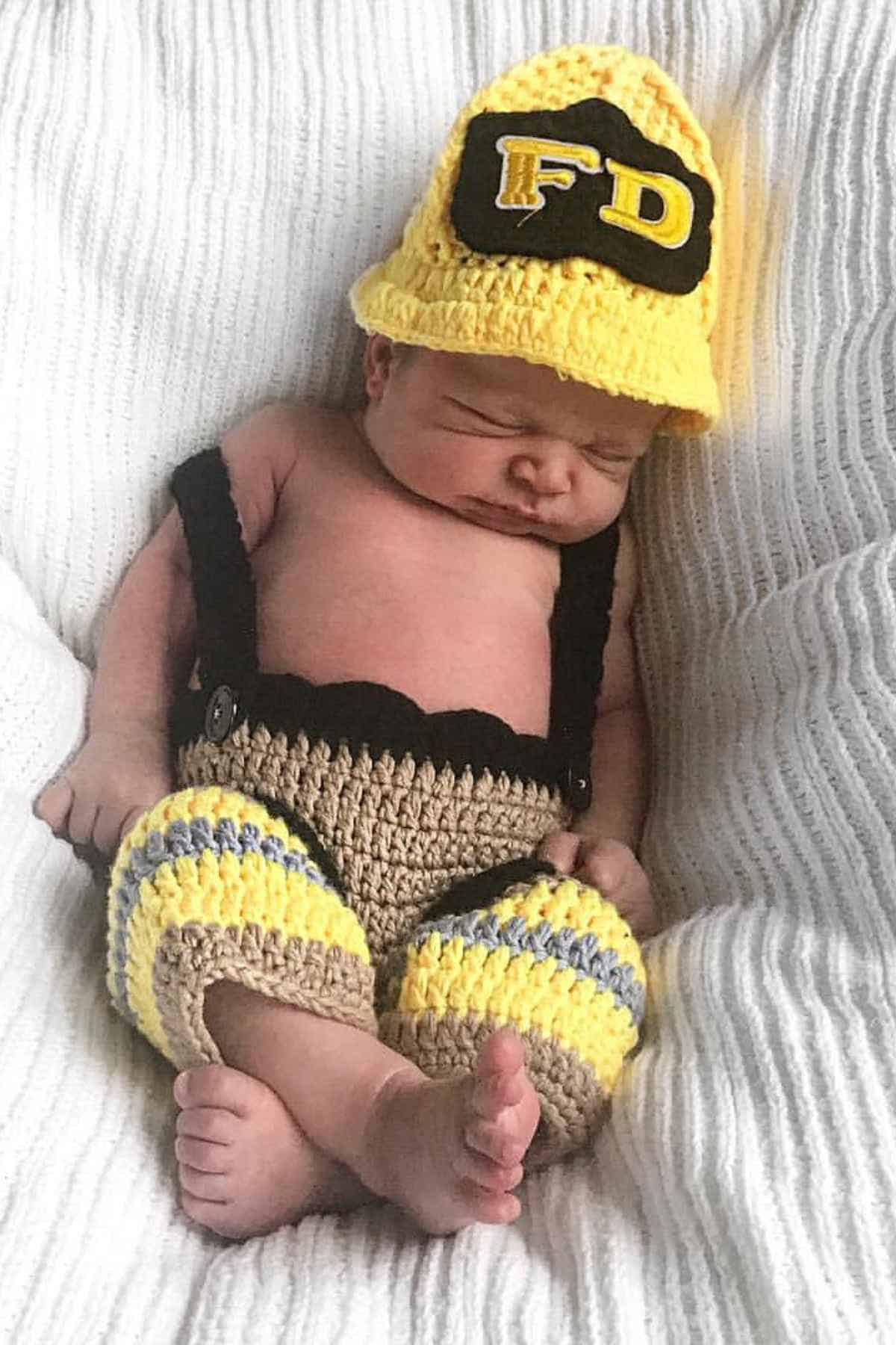 A baby boy in bed wearing a yellow, and brown knitted costume paired with yellow hat with "FD" text imitating a fire fighter.