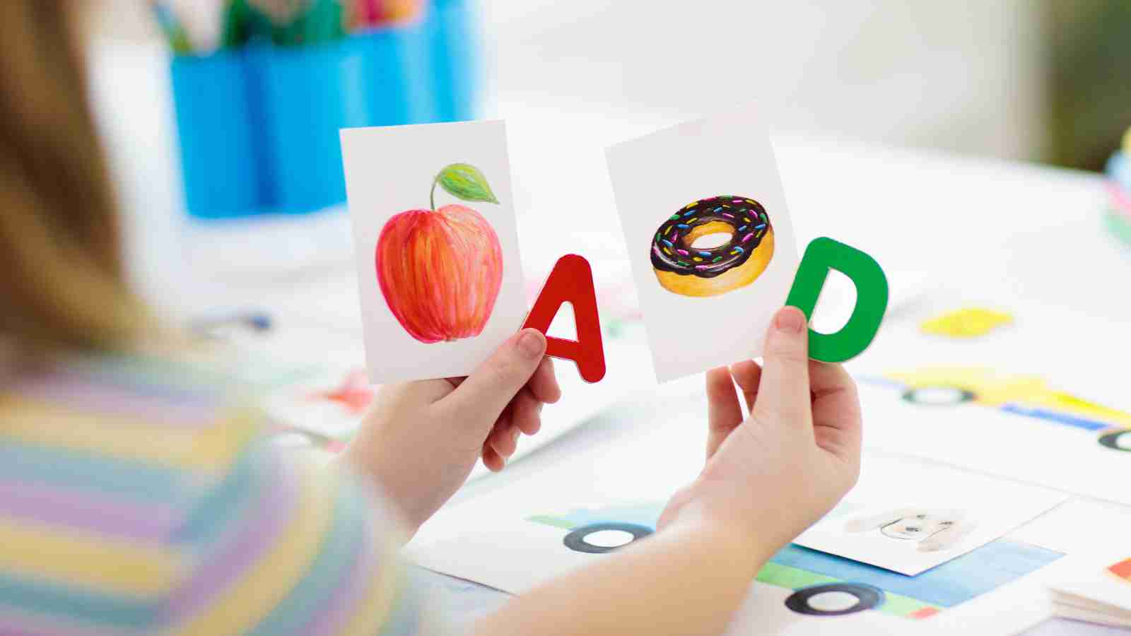 A child holds two cards, one with an apple and the letter A, and another with a donut and the letter D.