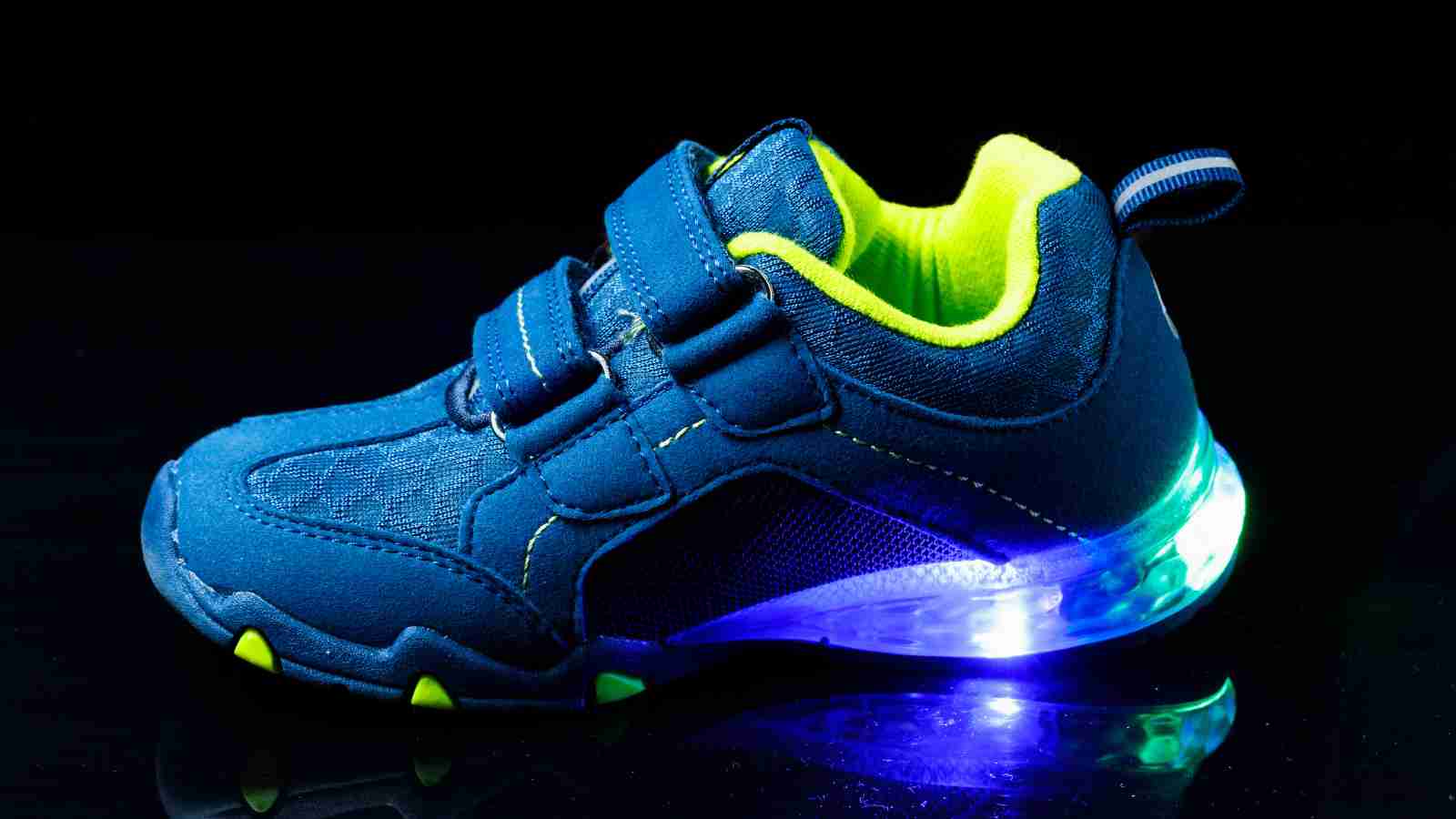 A blue children's sneaker with neon yellow accents and a glowing light-up sole, photographed against a black background.