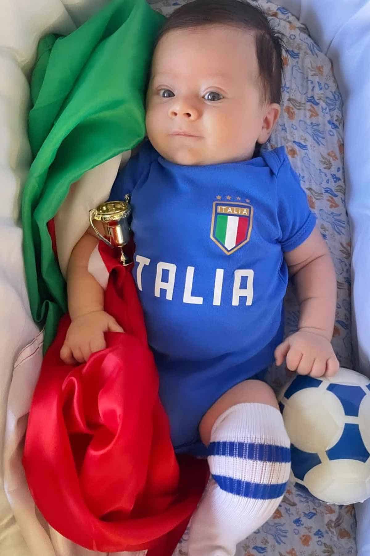 A baby boy dressed in an blue Italian football jersey, with a ball and some green and red cloth beside him.